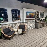 A patio setup featuring two wicker chairs with black cushions, a small table, plants, and a grill against a wall with stone and shiplap siding. A framed TV displays a mountain scene.