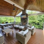 A custom hillside porch in Glen Mills features a spacious outdoor patio with wicker furniture, a wooden ceiling, a ceiling fan, and a stone fireplace with a TV mounted above, all surrounded by lush greenery.