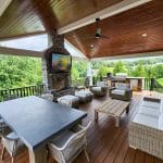 Custom hillside porch in Glen Mills featuring wicker furniture, a long dining table, cushioned chairs, a fireplace, and a TV. The area is covered with a wooden ceiling and overlooks a lush green landscape.