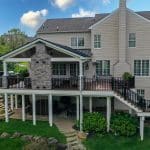 A two-story house with beige siding features a wooden deck with a stone fireplace and railing, supported by white pillars. The backyard has green grass, bushes, and a stone walkway.