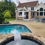 A suburban home with a partially stone exterior has a backyard featuring a swimming pool, patio area, and a hot tub in the foreground. The yard includes greenery and a fence in the background.