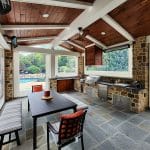 Outdoor kitchen and dining area with stone walls and a wooden ceiling, equipped with a grill, countertops, and seating, overlooking a yard with a pool.