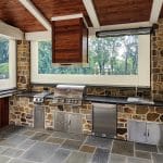 An outdoor kitchen with stone walls featuring a built-in grill, a stainless steel sink, and various storage cabinets beneath a wooden ceiling.