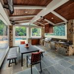 Outdoor kitchen with stone flooring, wooden ceiling, and built-in appliances. A table with two chairs and a bench sits in the center. A view of greenery and a pool is visible in the background.