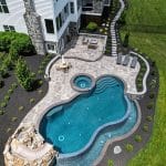 Aerial view of a backyard featuring a stone patio with a swimming pool, hot tub, water slide, lounge chairs, and a small grassy area, adjacent to a multi-story house.