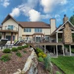 A large house with beige siding features multiple balconies, a custom deck in Reading with outdoor seating, a stone chimney, and well-maintained landscaping with a lawn and garden.