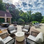 Outdoor patio with chairs and a small table near a swimming pool. A dining table with an umbrella and a grill are on a wooden deck. Trees and a cloudy sky are in the background.