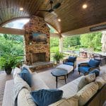 A covered outdoor patio with a stone fireplace, ceiling fan, cushioned seating, and a dining area, overlooks a green garden with a pool in the background.