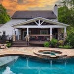 Backyard view of a house with a wooden deck, covered patio, stone detailing, and a swimming pool with an integrated hot tub at sunset. Trees surround the property, creating a secluded atmosphere.