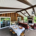 An outdoor patio with a dining table set up for six people, a seating area with a sofa and chairs, stone fireplace, wooden ceiling beams, and a view of greenery outside.