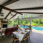 Covered outdoor patio with a dining table set for a meal, cushioned seating, a fireplace, television, and a view of a pool surrounded by greenery. A pink flamingo float is in the pool.