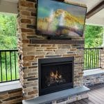 Outdoor patio with a stone fireplace and a mounted TV displaying a mountain scene. The area has a partially covered roof and is surrounded by greenery.