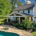 A two-story white house with a covered patio overlooks a stone-paved backyard with a pool and hot tub, surrounded by trees and greenery.