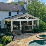 A two-story house with a spacious backyard featuring a covered patio, stone steps, and a pool with an adjacent hot tub, surrounded by trees and greenery.