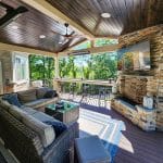 A covered outdoor patio with wicker furniture, a coffee table, a rug, a wall-mounted TV above a fireplace, and a ceiling fan under a wooden roof. Trees and greenery are visible in the background.