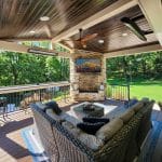 Covered outdoor patio with wicker furniture, cushioned seating, a coffee table, ceiling fan, and a mounted picture. The patio overlooks a lush green lawn and trees in the background.