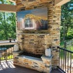 Outdoor patio with a stone fireplace, a mounted television, and decorative lanterns. There is also a bar-style table with stools and a view of a forested area in the background.