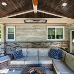 Outdoor patio with a wooden ceiling, wicker sectional sofa with cushions and pillows, and a stone wall with two windows. A rectangular heater is mounted on the wall. A table holds a book and remote.