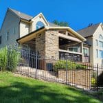 Two-story beige house with a peaked roof, stone chimney, and fenced entrance. Front lawn has green grass and shrubs. Clear blue sky in the background.