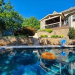 A backyard pool with a yellow inflatable ring, adjacent to a house with a stone exterior, a balcony, stairs, and lush green trees in the background.