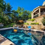 A backyard features a swimming pool with lounge chairs around it, surrounded by trees and greenery. A stone house with a deck overlooks the pool. Various pool toys are floating in the water.