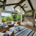 A covered outdoor patio with wicker furniture, a striped rug, and a TV mounted above a stone fireplace. Ceiling fans and heaters are visible. The area overlooks a lush green landscape.