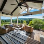 A covered outdoor patio features wicker furniture, a striped rug, a ceiling fan, and a TV mounted near a stone wall, overlooking a scenic view of grassy hills and trees.