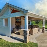 A covered outdoor pavilion with patio furniture, a TV mounted above a fireplace, and a bar area with stools, set on a paved area surrounded by grass and trees.