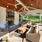 Covered outdoor patio with stone fireplace, mounted TV, ceiling fan, rattan furniture, and a wooden coffee table. Kitchen area with a grill is visible in the background.