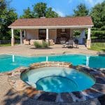 A backyard pool and spa with a stone border, adjacent to a furnished shed-like structure with a covered patio and a small kitchen setup. Trees and outdoor lounge chairs are in the background.