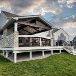 A modern house with an elevated patio featuring stone accents, a slanted roof, and outdoor seating, surrounded by a green lawn under a partly cloudy sky.