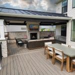 A modern outdoor patio with a covered seating area, fireplace, and mounted TV. Adjacent is a dining table with six chairs, set against a white house with large windows and sliding doors.