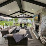 Outdoor covered patio with a seating area, fireplace, mounted TV, and wooden beams. Stone accents and a large rug enhance the cozy setting. Green lawns and houses are visible in the background.