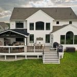 A two-story white house with a large backyard deck featuring a covered seating area, grill, and steps leading down to a grassy lawn. The sky is cloudy, and other houses are visible in the background.