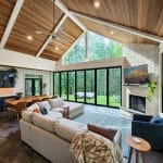 A modern living room with vaulted wooden ceilings, large windows, a sectional sofa, TV above the fireplace, built-in bar area, and dining table, overlooking a lush green backyard.