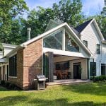 A modern two-story house with white siding, large windows, and a brick accent wall. The home features a partially open sliding glass door leading to a patio with a grill, surrounded by grass and trees.