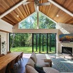 Spacious living room with a high wooden ceiling, large folding glass doors overlooking a green yard, a sectional sofa, a long dining table, a fireplace, and a wall-mounted TV.