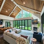 A spacious living room with a high wooden ceiling, large windows, a grey sectional sofa, a round side table, and a TV mounted above a fireplace. The room opens to a view of greenery outside.