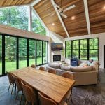 Modern living room with large windows overlooking greenery. Features a wooden dining table, cozy seating area with a fireplace, and a vaulted wood-paneled ceiling with recessed lighting.