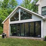 A modern house with white siding and large glass windows featuring a spacious patio area with a barbecue grill and surrounded by green trees and a manicured lawn.