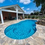 A backyard with a swimming pool, covered patio area, and a seating area surrounded by a brick wall and foliage.