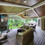 Covered patio with wooden ceiling, wicker furniture, a stone fireplace, wall-mounted TV, ceiling fan, and view of a garden through railings. The area includes a door leading inside the house.