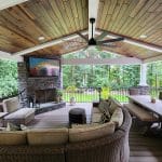 A covered outdoor living space with a cushioned sectional sofa set, a dining table with wicker chairs, a mounted TV above a stone fireplace, and a ceiling fan under a wood-paneled roof.