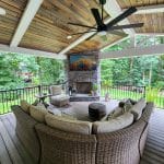 Covered patio with a ceiling fan, curved wicker sofa, cushioned chairs, round ottoman, and a fireplace. A TV is mounted above the fireplace, and the area is surrounded by lush green trees.