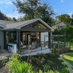 Modern backyard with a covered patio area, outdoor seating, a well-maintained garden, and a small putting green. Clear blue sky and trees in the background.