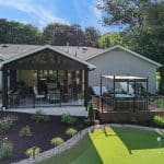 A backyard featuring a covered patio with seating and an outdoor deck with furniture alongside a well-maintained putting green on a sunny day.