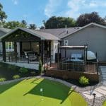 A backyard with a putting green, a deck with outdoor furniture and a grill, and a covered patio attached to a gray house with large windows and decorative plants.