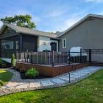 Modern backyard with a wooden deck, black railings, and a covered outdoor seating area attached to a house. Pathway with stepping stones leads to the deck. Lawn and landscaping surround the area.