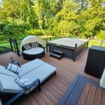 A modern outdoor deck with a cushioned lounge chair, a round daybed with a canopy, a hot tub, and greenery in the background.