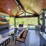 A modern outdoor living room features wooden ceiling, a fireplace, a TV, cushioned seating, large windows with outdoor views, and multiple light fixtures.